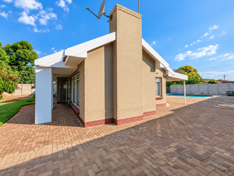 Rear view of the house featuring a paved patio area, covered veranda, brick chimney, and a swimming pool set within a fenced garden.