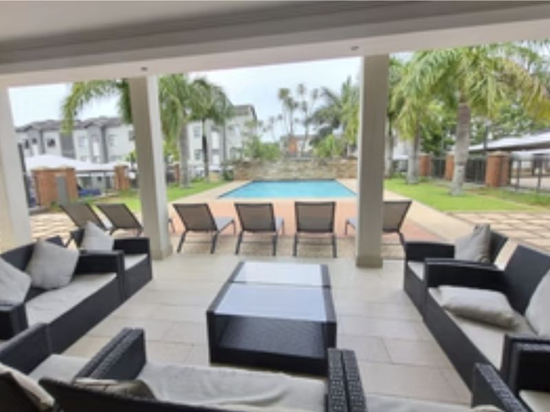 A spacious covered patio area overlooking a communal swimming pool and landscaped garden, featuring tiled flooring and modern structural columns.