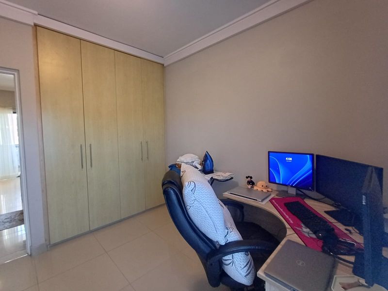 A functional bedroom featuring light-toned built-in wardrobes, neutral tiled flooring, and decorative cove cornicing, currently utilized as a home office space.