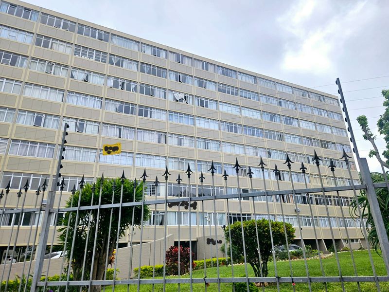 Exterior view of a multi-story apartment building featuring a metal security fence with electric wiring and large windows.