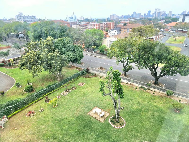 Elevated view overlooking a well-maintained communal garden area with mature trees, perimeter fencing, and surrounding urban neighborhood infrastructure.