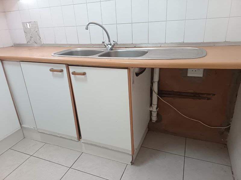 A dated kitchen area featuring a double bowl sink, tiled splashback, and base cabinetry with visible wear and exposed plumbing requiring maintenance.