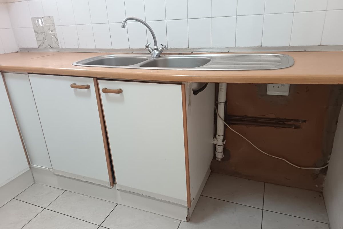 A dated kitchen area featuring a double bowl sink, tiled splashback, and base cabinetry with visible wear and exposed plumbing requiring maintenance.