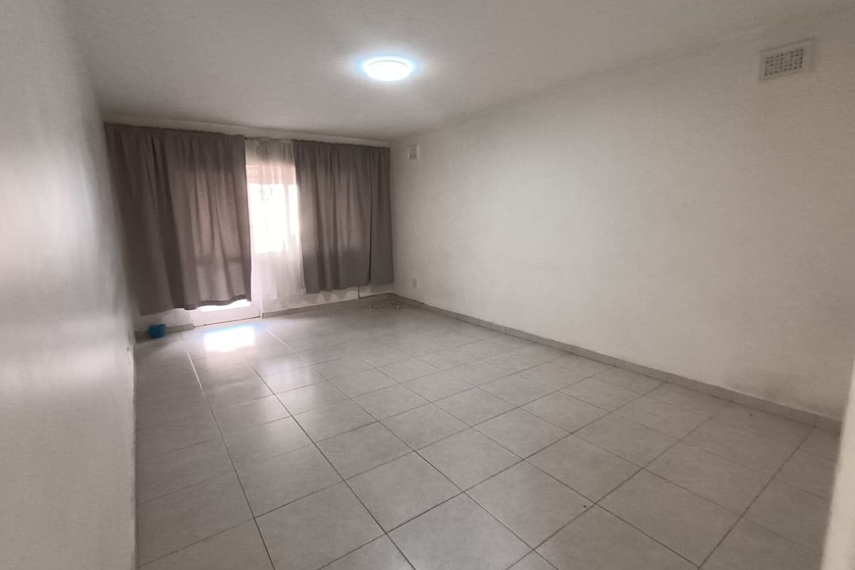 An empty, neutrally finished living room featuring tiled flooring and natural light access through a window with curtain tracks.