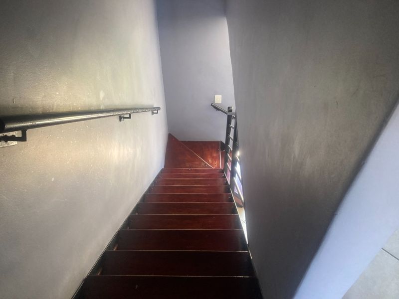 A narrow interior staircase featuring dark wooden treads, a metal handrail, and a metal balustrade, showing signs of wear on the walls and steps.