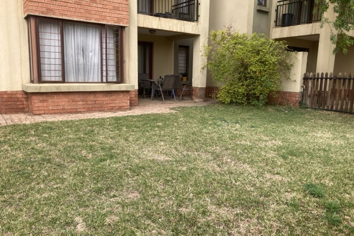 Rear view of the apartment building featuring a private garden, paved patio area, and covered balcony with metal railings.
