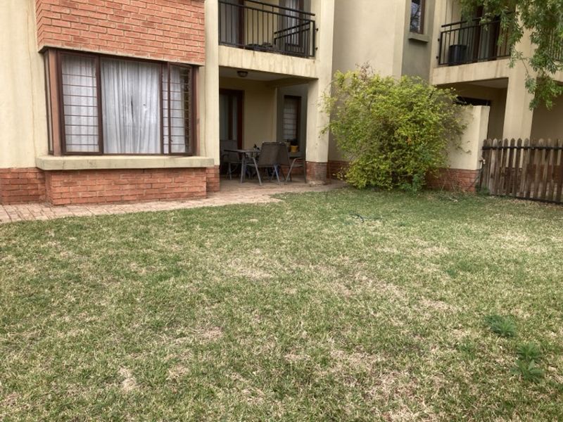 Rear view of the apartment building featuring a private garden, paved patio area, and covered balcony with metal railings.