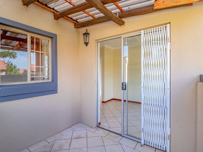 A covered patio area featuring tiled flooring, a timber pergola, and a sliding glass door with a security gate providing access to the interior.