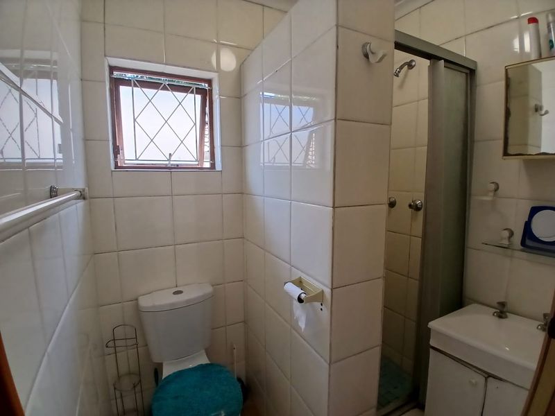 A functional bathroom featuring a shower enclosure, toilet, and vanity basin with tiled walls and a window with security bars.