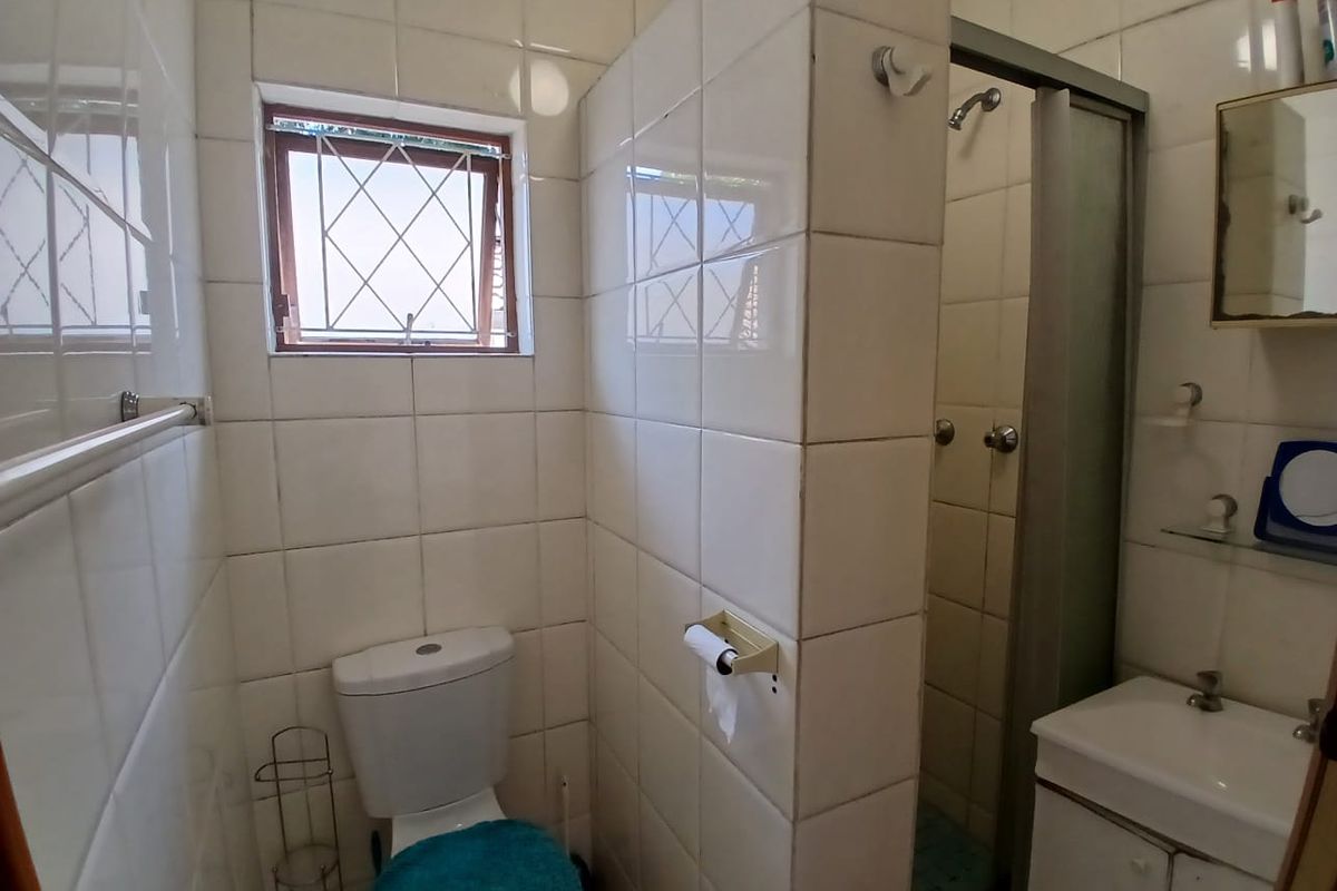 A functional bathroom featuring a shower enclosure, toilet, and vanity basin with tiled walls and a window with security bars.