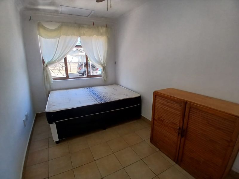 A simple bedroom featuring tiled flooring, a ceiling fan, and a window fitted with security bars.