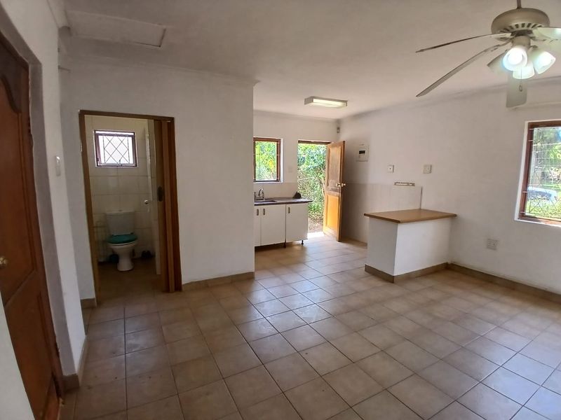 Open-plan living area featuring tiled floors, a ceiling fan, a compact kitchen with built-in cabinetry, and an adjacent bathroom.