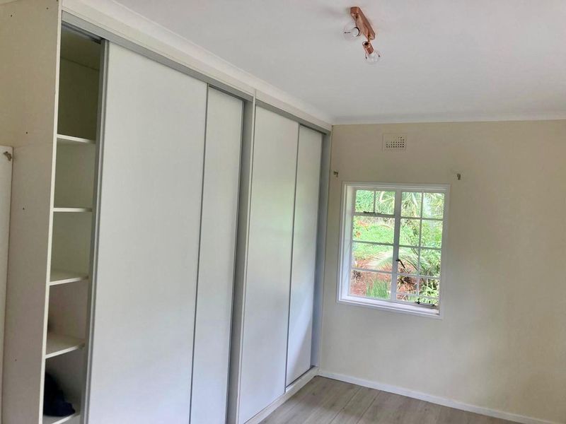 A bright, empty bedroom featuring built-in sliding door wardrobes, laminate flooring, and a window overlooking the garden.