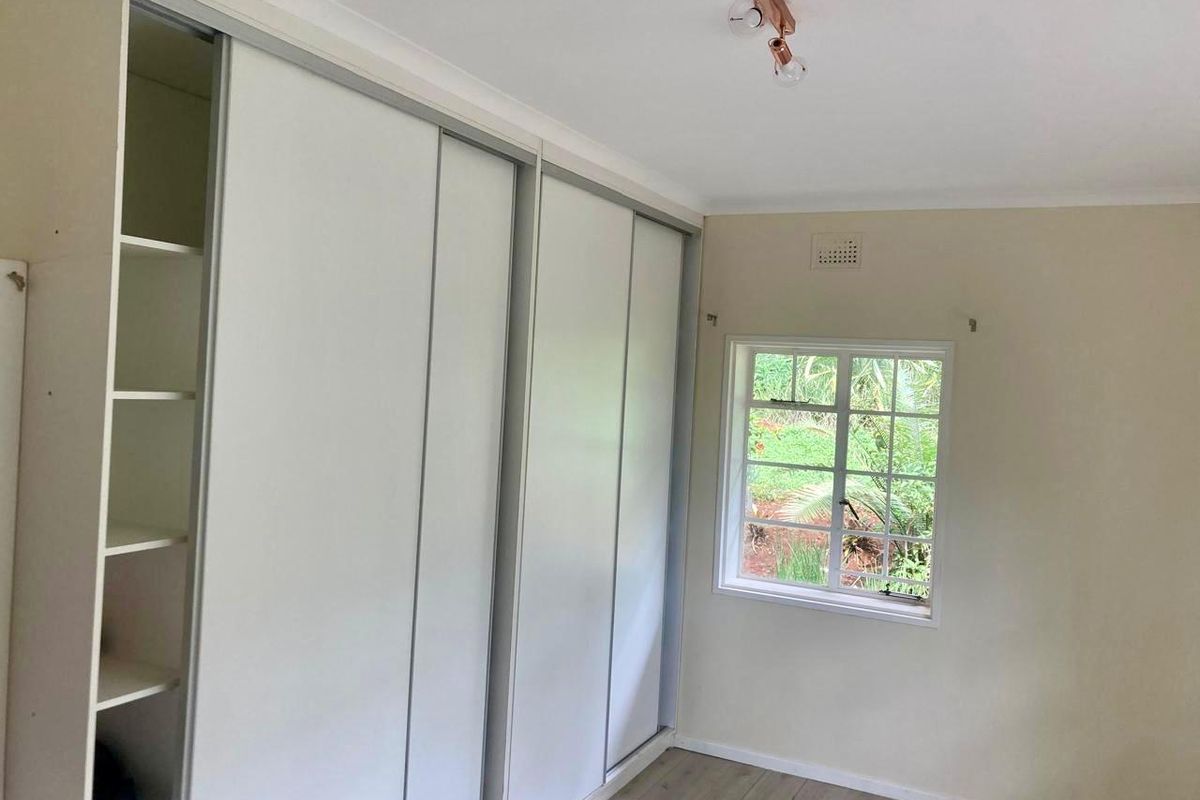 A bright, empty bedroom featuring built-in sliding door wardrobes, laminate flooring, and a window overlooking the garden.