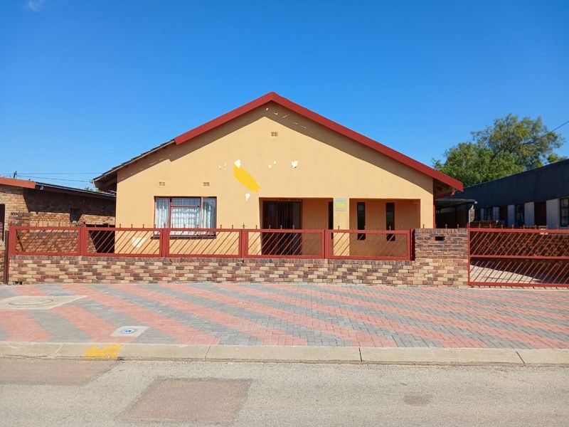 Front elevation of a single-story house featuring a brick boundary wall, metal security fencing, and a paved driveway area.