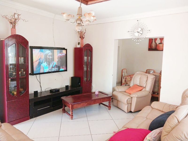 A comfortable living room featuring tiled flooring, decorative ceiling cornices, and a central chandelier, with an open-plan flow into the adjacent dining area.