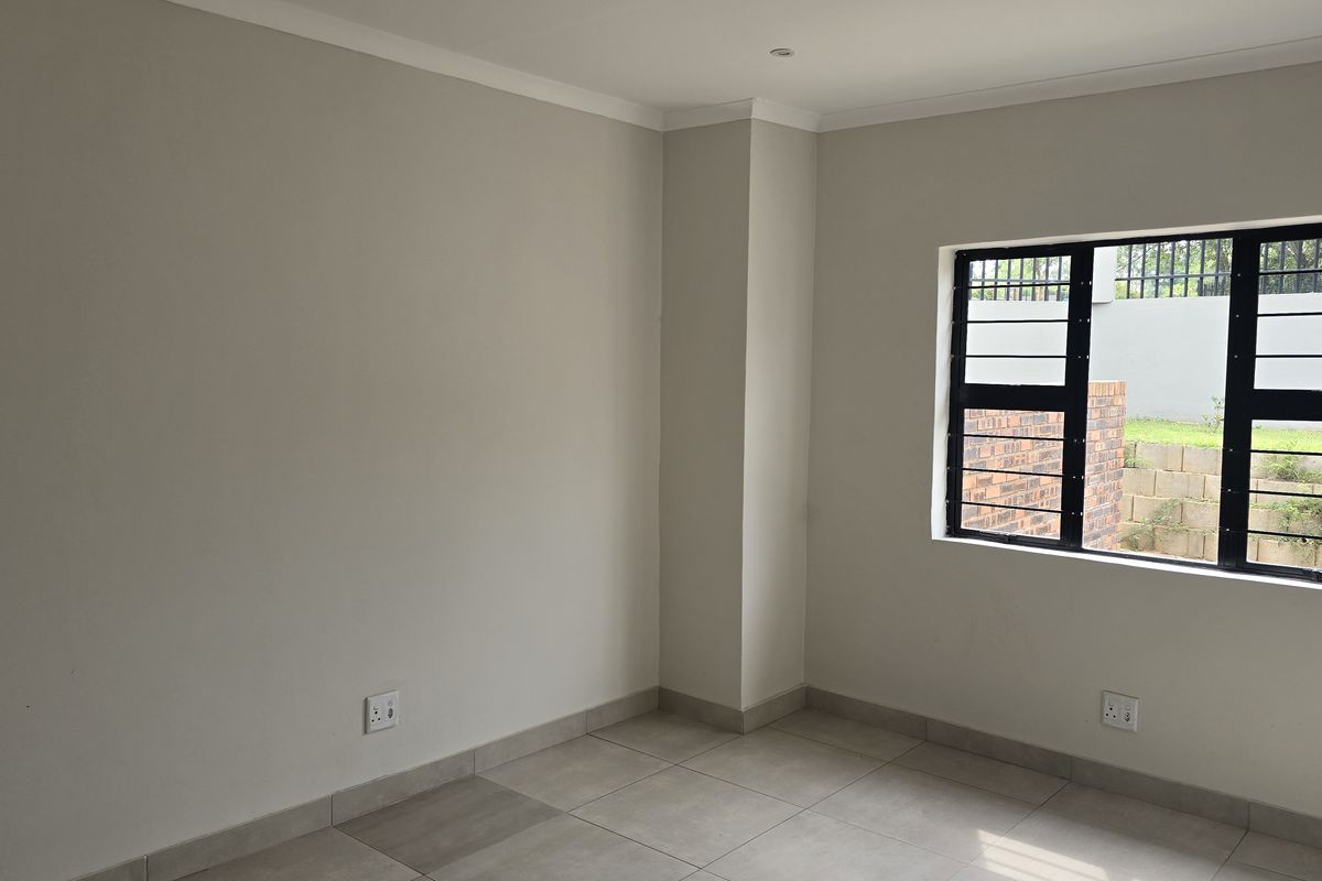 A clean, empty bedroom featuring neutral wall tones, tiled flooring, and large windows with security bars, offering a bright and versatile living space.