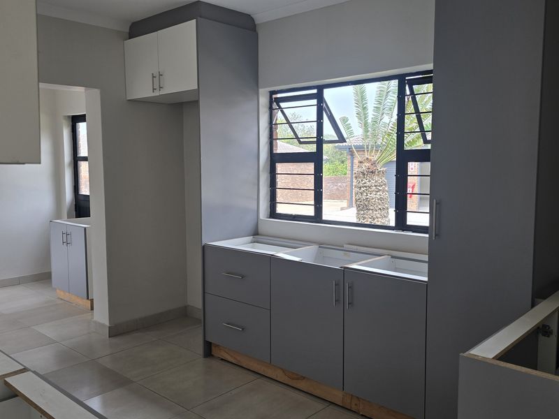 A kitchen space currently undergoing renovation, featuring grey built-in cabinetry, tiled flooring, and large windows with views of the exterior.