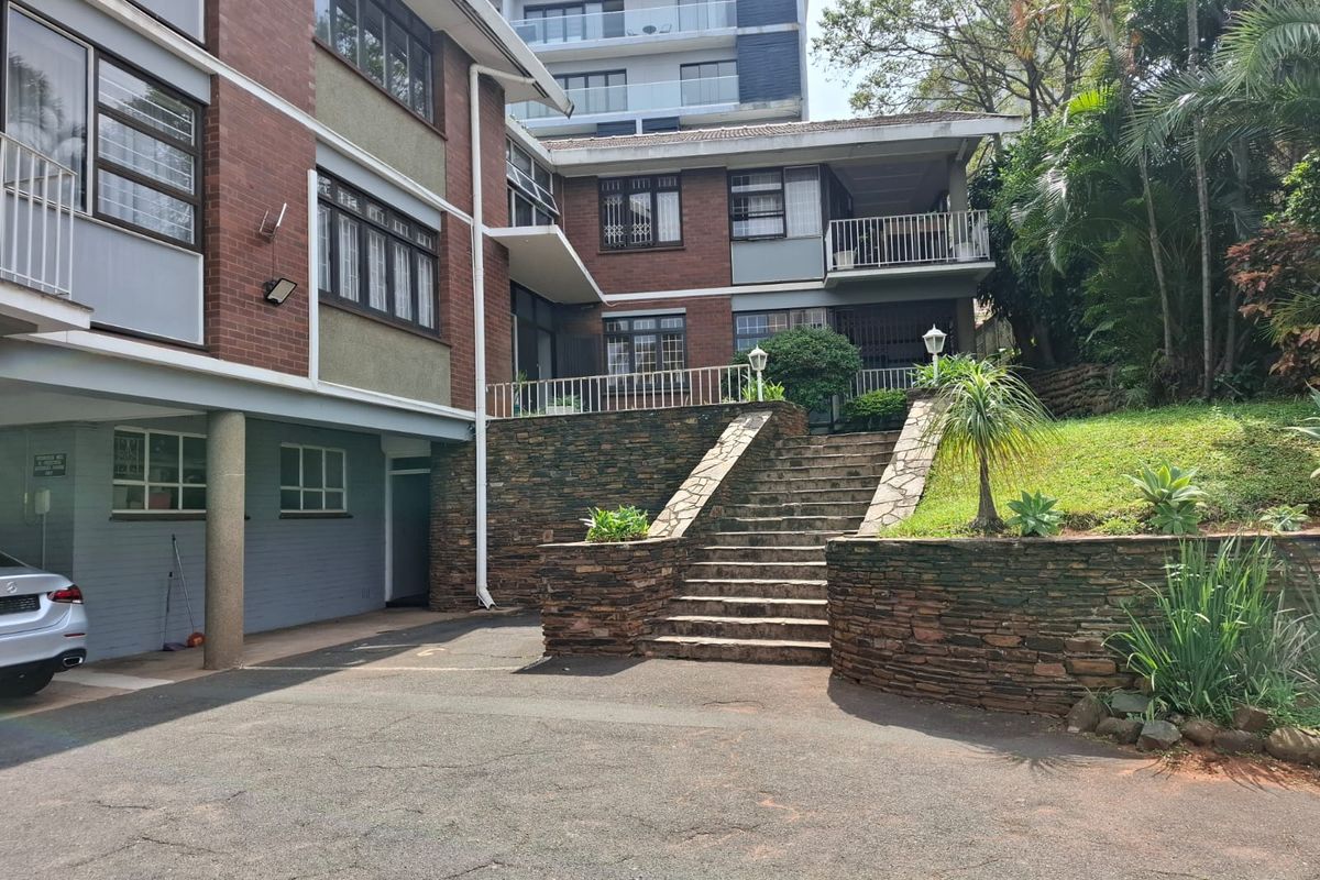 Rear exterior view of the flat complex featuring a brick facade, stone retaining walls, a central staircase, and a balcony with metal railings.