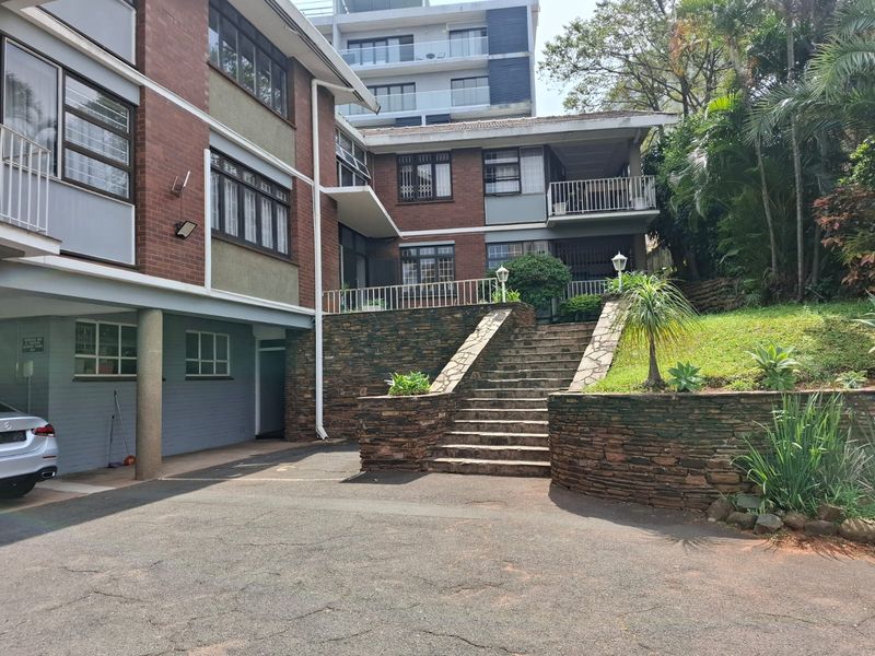 Rear exterior view of the flat complex featuring a brick facade, stone retaining walls, a central staircase, and a balcony with metal railings.