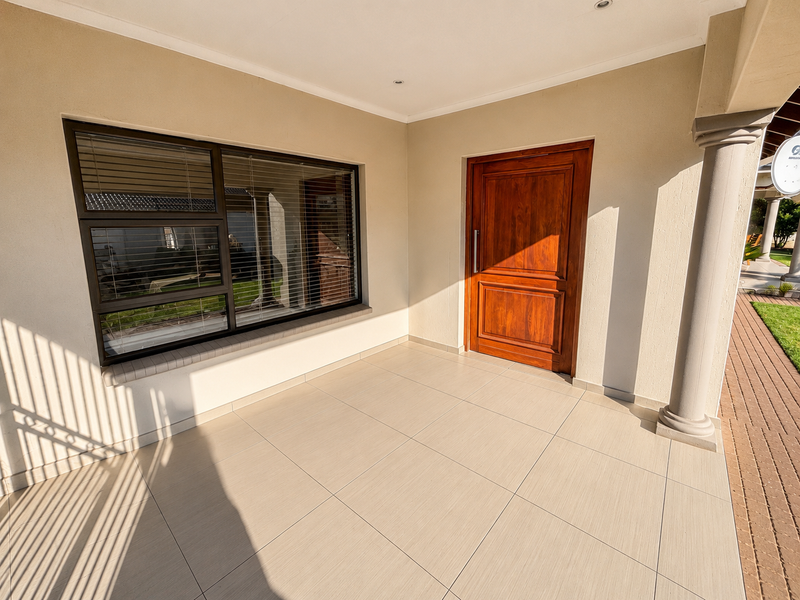 A clean, covered patio area featuring large format floor tiles, a solid wooden entrance door, and modern aluminium window frames.
