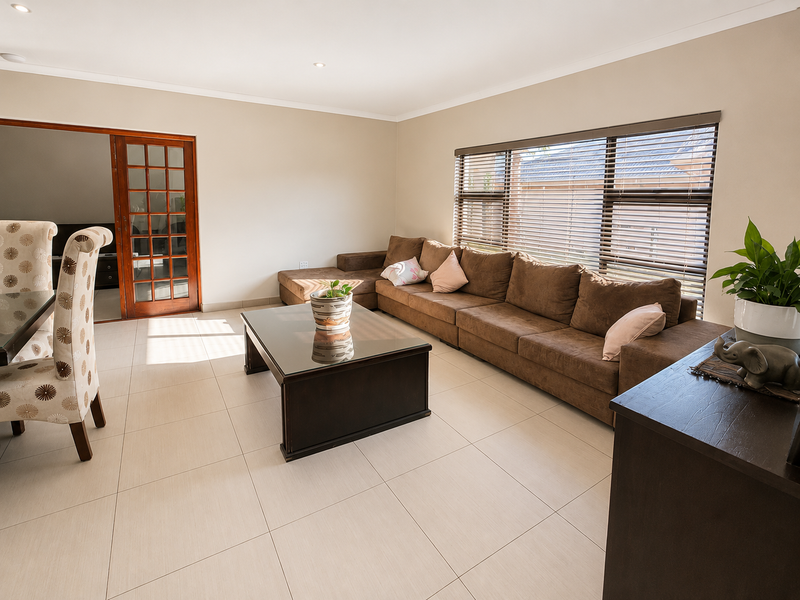 A bright and spacious living room featuring neutral tiled flooring, recessed downlighting, and large windows fitted with venetian blinds, leading to an adjacent room.