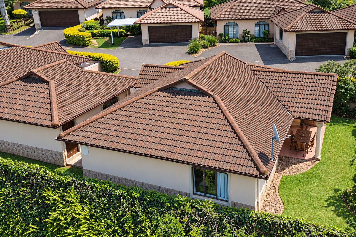 An aerial view of a townhouse complex featuring tiled roofs, paved driveways, garages, and private garden spaces with covered patios.