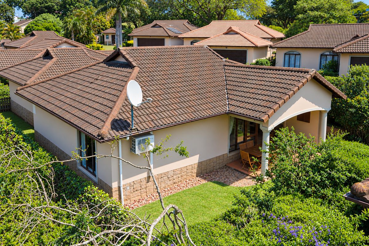 An elevated view of the rear exterior of a townhouse, featuring a tiled roof, covered patio area, and established garden surroundings.