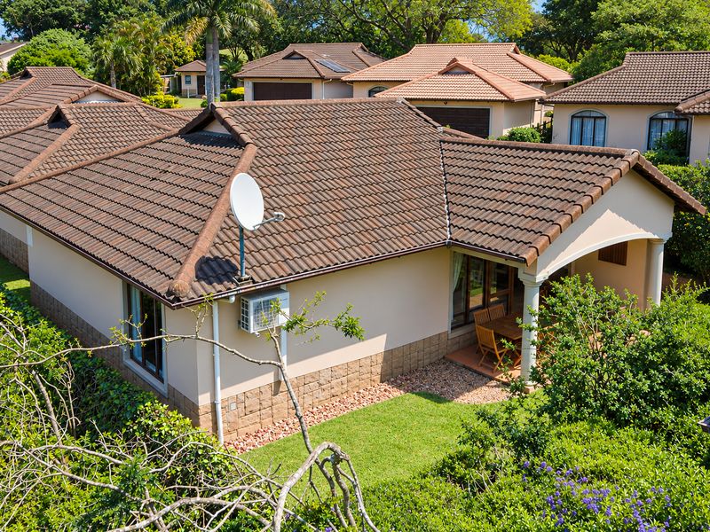 An elevated view of the rear exterior of a townhouse, featuring a tiled roof, covered patio area, and established garden surroundings.