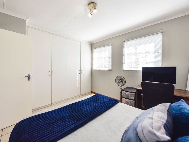 A well-lit bedroom featuring built-in cupboards, tiled flooring, and dual windows with venetian blinds, offering a clean and functional living space.