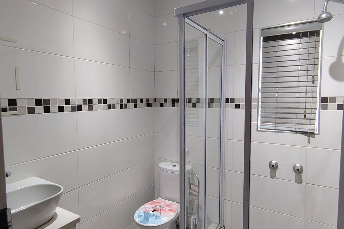 A clean, modern bathroom featuring a glass-enclosed shower, vanity basin, and toilet with neutral tiled finishes and a window for natural light.