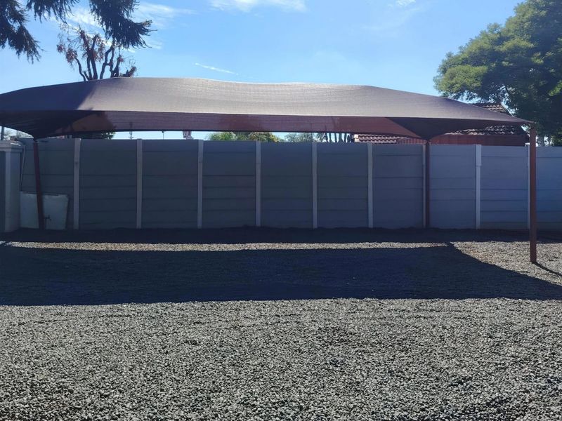 A gravel driveway featuring a large shade cloth carport structure and a perimeter precast concrete boundary wall.