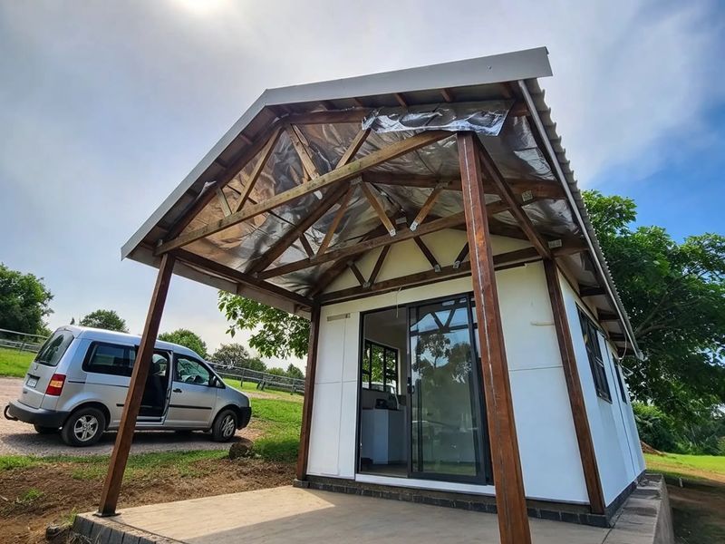 Exterior view of a cottage featuring a raised concrete patio, timber roof structure with exposed insulation, and a sliding glass entrance door.