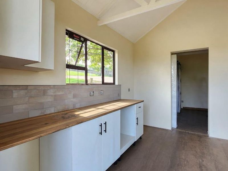 A bright, modern kitchen area featuring white cabinetry, a warm timber countertop, subway tile backsplash, and a high vaulted ceiling with exposed trusses.