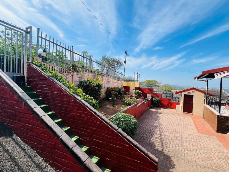 A paved rear courtyard featuring tiered brick retaining walls, landscaped garden beds, metal security fencing, and a small detached outbuilding.