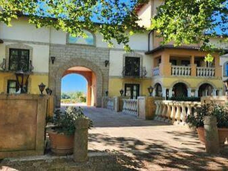 A Mediterranean-style apartment complex entrance featuring a prominent stone archway, decorative balustrades, and a paved driveway approach.