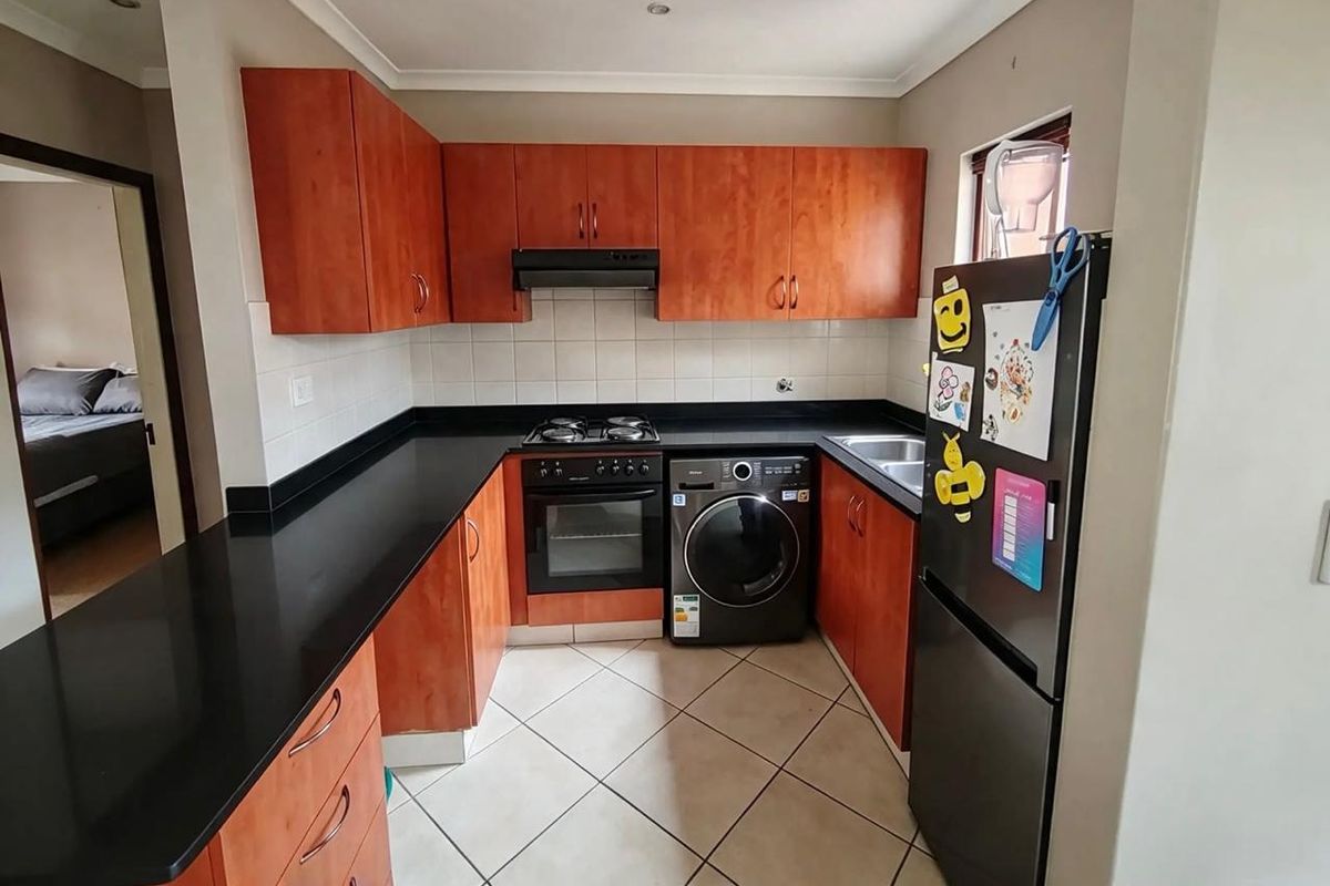 A functional kitchen featuring wood-finish cabinetry, black countertops, a built-in oven with gas hob, and tiled flooring with integrated laundry appliance space.