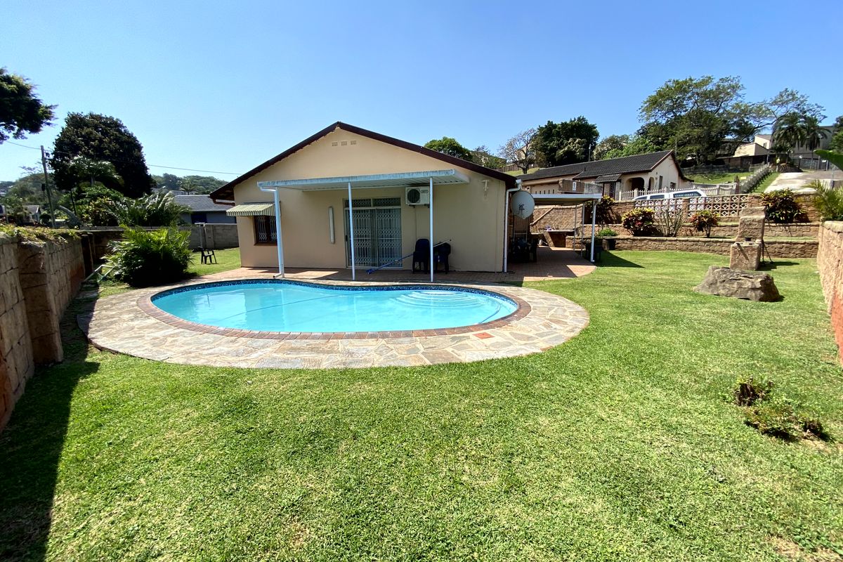Rear view of the property featuring a swimming pool with stone paving, a covered patio area, and a well-maintained garden with retaining walls.