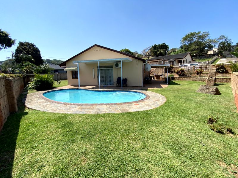 Rear view of the property featuring a swimming pool with stone paving, a covered patio area, and a well-maintained garden with retaining walls.