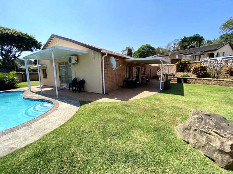 Rear exterior view featuring a swimming pool, paved patio area with covered roofing, and a landscaped garden with retaining walls.