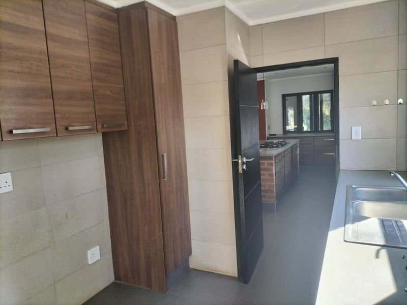 A clean, modern scullery featuring wood-finish cabinetry, tiled walls, and a double sink, with a view through to the main kitchen area.