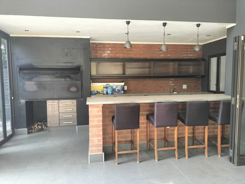 Covered patio area featuring a built-in braai, brick feature wall, stone-topped bar counter with sink, and modern pendant lighting.