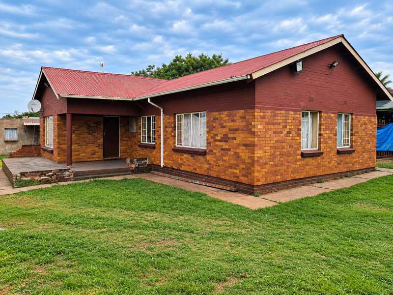 A single-story brick house featuring a red corrugated metal roof, a covered front porch, and a surrounding lawn area.