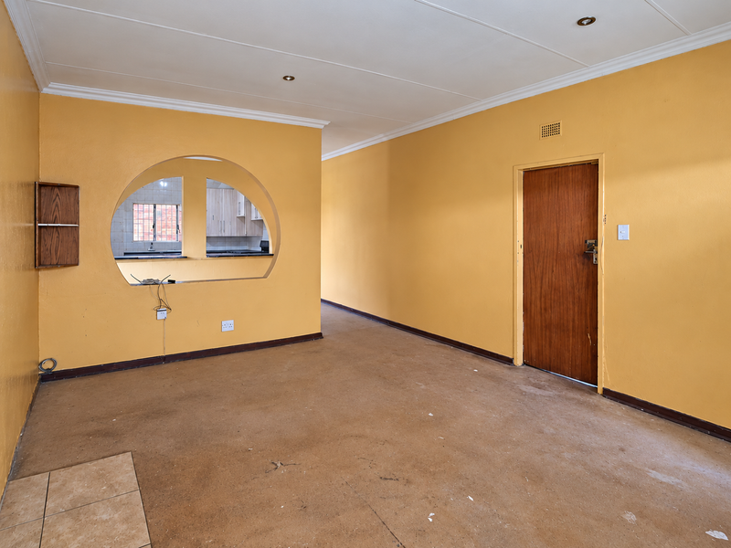 An empty living room featuring a distinctive arched opening to the kitchen, recessed lighting, and a wooden door, requiring cosmetic updates to flooring and walls.