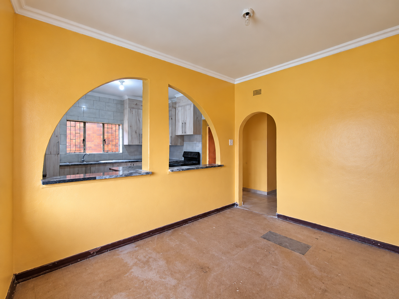 An empty living room featuring distinctive arched pass-through windows into the kitchen, coved ceilings, and original flooring, offering a blank canvas for renovation.