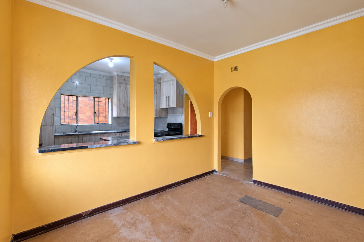 An empty living room featuring distinctive arched pass-through windows into the kitchen, coved ceilings, and original flooring, offering a blank canvas for renovation.