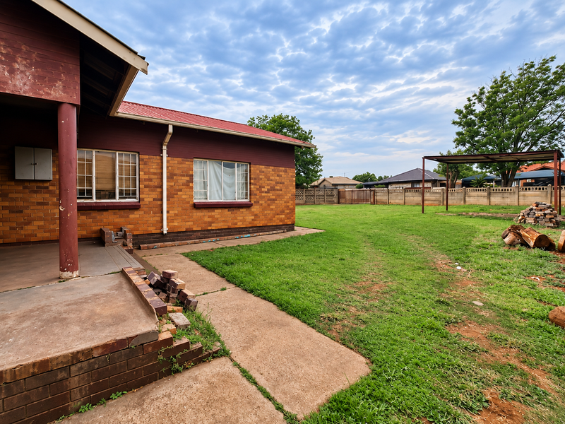 Rear view of the property featuring a brick facade, covered patio area, and a spacious backyard with a metal carport structure and perimeter walling.