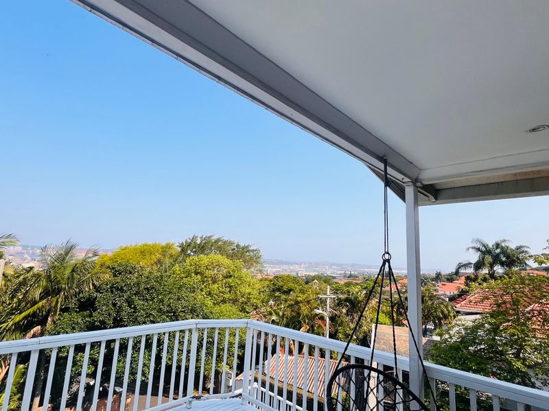 A spacious covered balcony featuring a white metal railing and elevated views over the surrounding neighborhood and lush greenery.