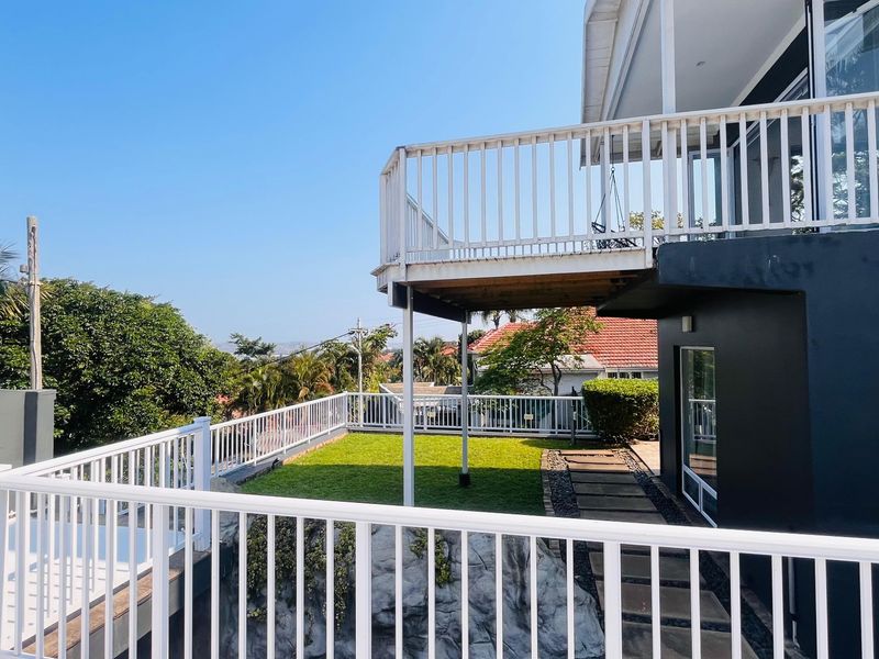 Rear view of the property featuring a raised balcony with white railings, a paved walkway, and a well-maintained lawn area.