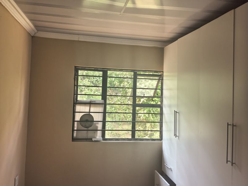 A bedroom featuring built-in cupboards, tiled flooring, and security bars on the window. The room is currently vacant and ready for personalization.